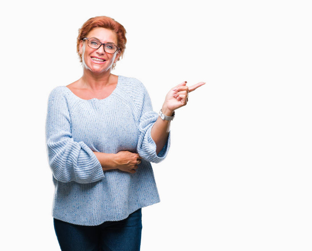 Atrractive vanhempi valkoihoinen punapää nainen silmälasit yli eristetty tausta iso hymy kasvoillaan, osoittaa käsin ja sormella sivuun katsoen kameraa
. - Valokuva, kuva
