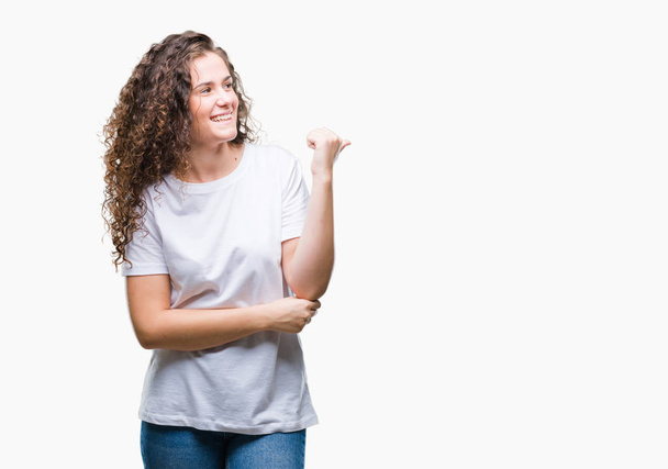 Beautiful brunette curly hair young girl wearing casual t-shirt over isolated background smiling with happy face looking and pointing to the side with thumb up. - Photo, Image