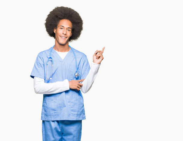 Giovane medico afro americano uomo con i capelli afro con un grande sorriso sul viso, che punta con mano e dito verso il lato guardando la fotocamera
. - Foto, immagini