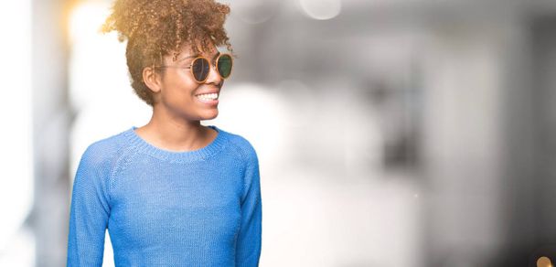 schöne junge afrikanisch-amerikanische Frau mit Sonnenbrille vor isoliertem Hintergrund, die mit einem Lächeln im Gesicht und natürlichem Ausdruck zur Seite schaut. Lachen selbstbewusst. - Foto, Bild
