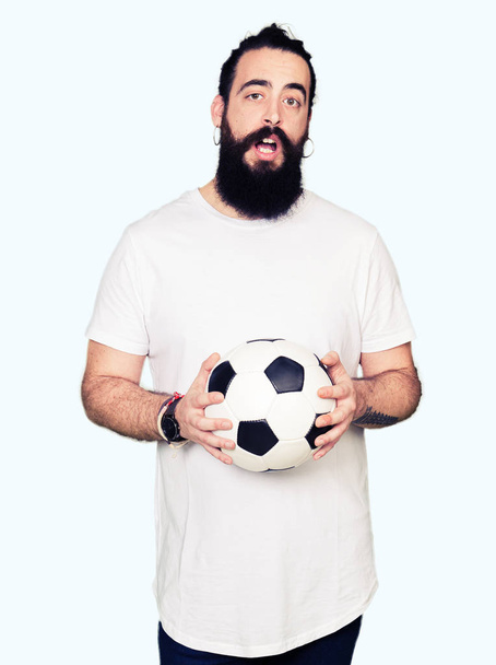 Young man with long hair and bear holding soccer football ball scared in shock with a surprise face, afraid and excited with fear expression - Foto, Imagen