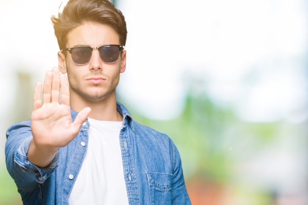 Jonge knappe man dragen zonnebril op geïsoleerde achtergrond stop zingen met de palm van de hand doen. Waarschuwing expressie met negatieve en ernstige gebaar op het gezicht. - Foto, afbeelding