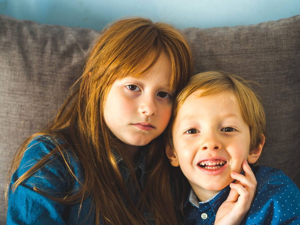 Dos niños hermanos rubios con camisas azules en un sofá abrazándose. Retrato de niños en interiores
 - Foto, Imagen