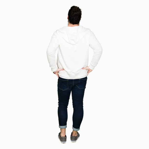 junger Hipster-Mann mit langen Haaren und Bart in sportlichem Sweatshirt, der rückwärts steht und mit den Armen am Körper wegschaut - Foto, Bild