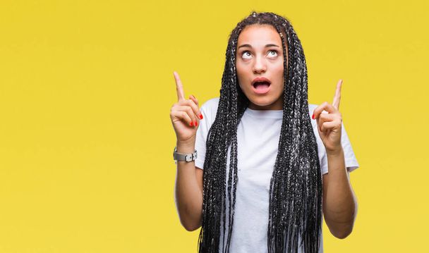 Young braided hair african american girl over isolated background amazed and surprised looking up and pointing with fingers and raised arms. - Photo, Image
