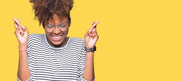 Linda jovem afro-americana vestindo óculos sobre fundo isolado sorrindo cruzando dedos com esperança e olhos fechados. Sorte e conceito supersticioso
. - Foto, Imagem