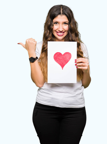 Young adult woman holding card with red heart pointing and showing with thumb up to the side with happy face smiling - Valokuva, kuva