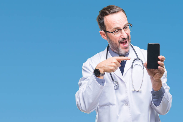 Middle age senior hoary doctor man showing smartphone screen over isolated background very happy pointing with hand and finger - Photo, Image
