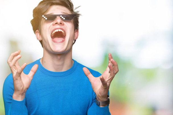 Young man wearing funny thug life glasses over isolated background crazy and mad shouting and yelling with aggressive expression and arms raised. Frustration concept. - Photo, Image