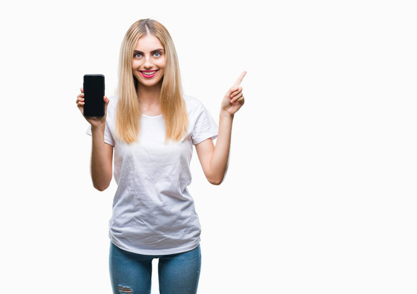 Joven hermosa mujer rubia mostrando teléfono inteligente sobre fondo aislado muy feliz señalando con la mano y el dedo a un lado
 - Foto, Imagen