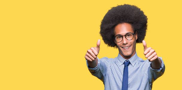 Jeune homme d'affaires afro-américain avec des cheveux afro portant des lunettes approuvant faire un geste positif avec la main, pouces levés souriant et heureux pour le succès. En regardant la caméra, geste gagnant
. - Photo, image