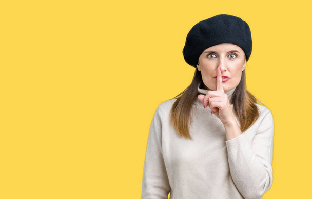 Middelbare leeftijd volwassen vrouw winter trui en baret dragen over geïsoleerde achtergrond vragen stil met de vinger op de lippen te zijn. Stilte en geheime concept. - Foto, afbeelding
