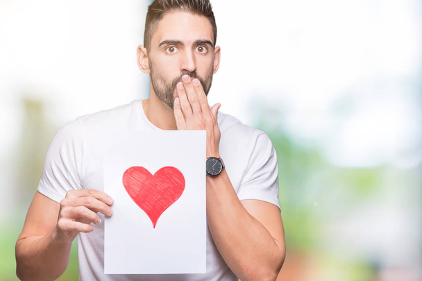 Handsome young man holding card with red heart over isolated background cover mouth with hand shocked with shame for mistake, expression of fear, scared in silence, secret concept - Photo, image