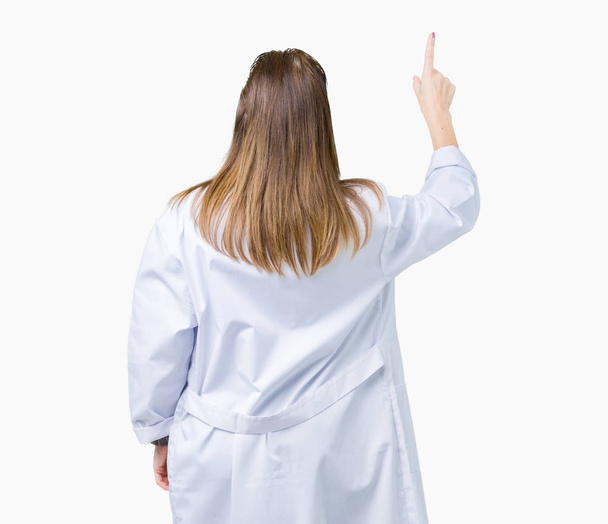 reife Ärztin mittleren Alters, die medizinischen Mantel über isoliertem Hintergrund trägt und rückwärts mit der Hand nach hinten zeigt - Foto, Bild
