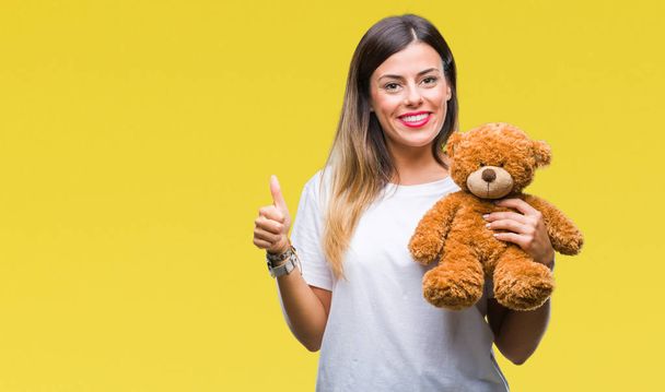 Young beautiful woman holding teddy bear plush over isolated background happy with big smile doing ok sign, thumb up with fingers, excellent sign - 写真・画像