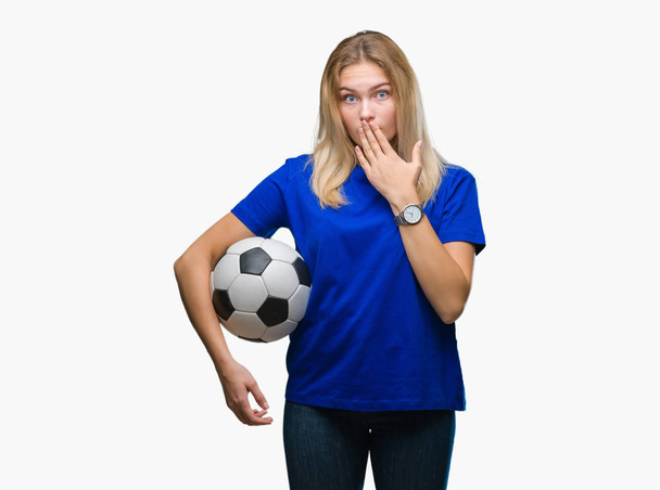Молодая кавказская женщина, держащая футбольный мяч на изолированном заднем плане, прикрывает рот рукой, потрясенной стыдом за ошибку, выражением страха, испуганной в тишине, секретной концепцией
 - Фото, изображение