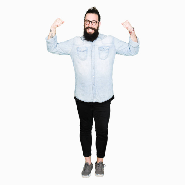 Young hipster man with long hair and beard wearing glasses showing arms muscles smiling proud. Fitness concept. - Φωτογραφία, εικόνα