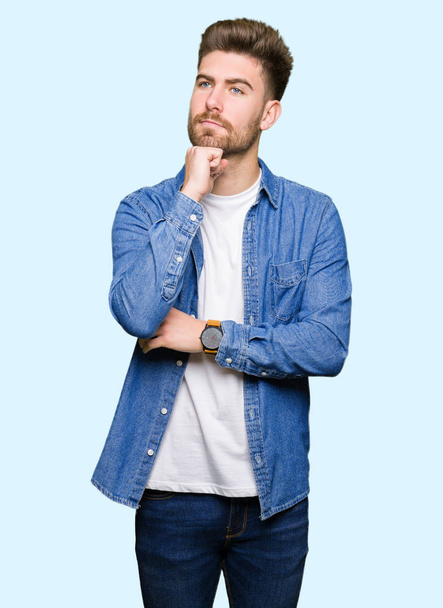 Young handsome blond man wearing casual denim jacket with hand on chin thinking about question, pensive expression. Smiling with thoughtful face. Doubt concept. - Foto, Imagen