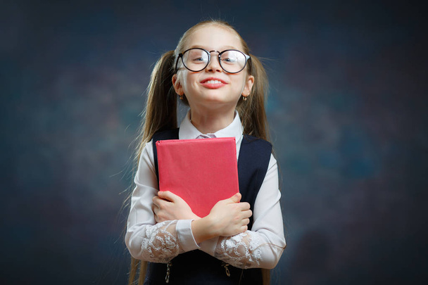 fröhliches kleines Schulmädchen in Uniform hält Buch fest. Nettes intelligentes Mädchen mit großer Brille und traditionellem Schwarz-Weiß-Outfit. Gebildete Vorschulerzieherinnen bekommen Wissen - Foto, Bild