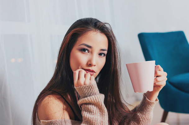 Candid portrait of sensual smiling asian girl young woman with dark long hair in cozy beige sweater holding cup of tea on white curtain background - 写真・画像