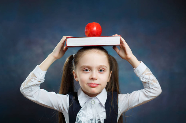 Симпатичная школьница держит книгу Apple на голове. Спокойная молодая девушка с двумя понюханными хвостами смотрит на камеру на темном фоне. Электронный школьный учебник без эмоций
 - Фото, изображение