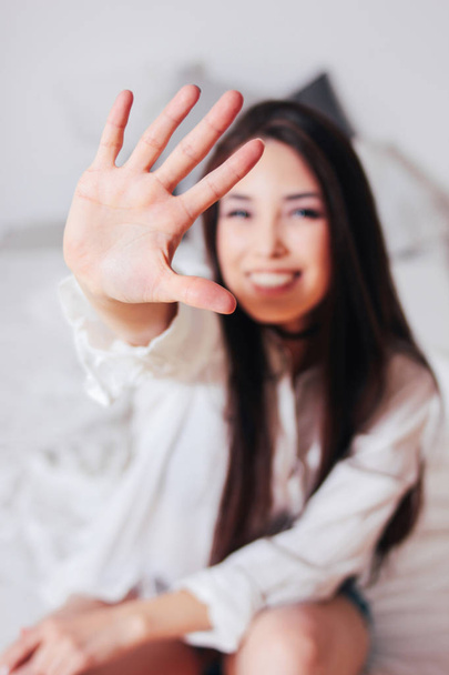 Szczęśliwy uśmiechający się piękne długie włosy Azjatyckie dziewczyna młoda kobieta wyciągając rękę w kierunku kamery siedzi na łóżku - Zdjęcie, obraz
