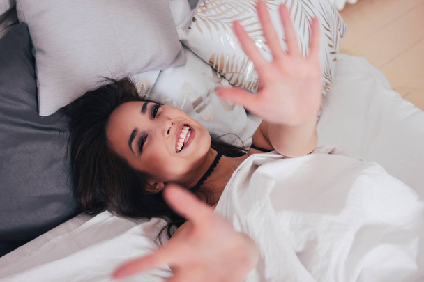 Feliz sonrisa hermosa pelo largo asiático chica joven acostada en la cama y tirando de las manos hacia la cámara, mañana acogedor
 - Foto, Imagen