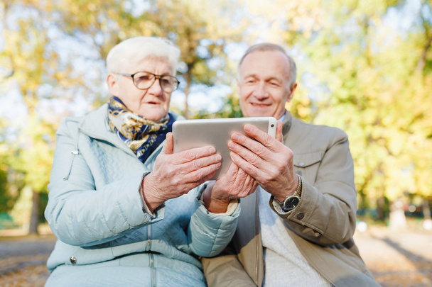 Senior para na ławce w parku jesienią i przy użyciu cyfrowego tabletu - Zdjęcie, obraz