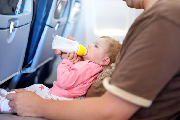 Father holding baby daughter during flight on airplane going on vacations. Baby girl drinking formula milk from bottle. Air travel with baby, child and family concept. Tired man traveling with kids. - Foto, Imagem