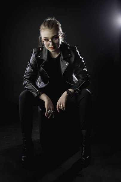 young girl in a black leather jacket and glasses on a dark backg - Фото, изображение