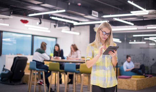 young blonde businesswoman working online using digital tablet while standing at modern startup office with multiethnic business team in background - Foto, immagini
