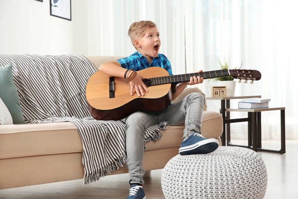 Cute little boy playing guitar on sofa in room - Zdjęcie, obraz