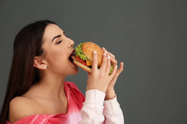 Νεαρή γυναίκα τρώει νόστιμο burger σε γκρι φόντο. Χώρο για το κείμενο - Φωτογραφία, εικόνα