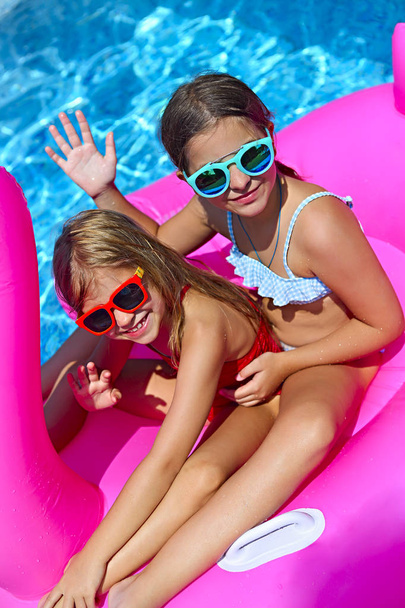 Portrait of two girls wearing sunglasses, happy friends on inflatable flamingo swim float, enjoying pool party in summer  - Photo, image