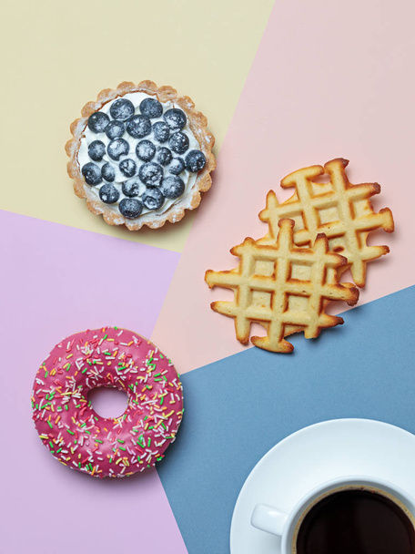 Плоский лежак з чорною кавою та смачними десертами пончик, чорничний пиріг та вафлі на фоні пастельного кольорового блоку
 - Фото, зображення
