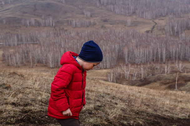 six-year-old boy in a red jacket walks outside the city in the spring in the highlands, the boy looks at his feet - Photo, image