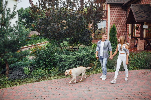 Moment of resting! Young attractive family walking the labrador near the house. Beautiful couple near modern home with dog on the leash. - 写真・画像