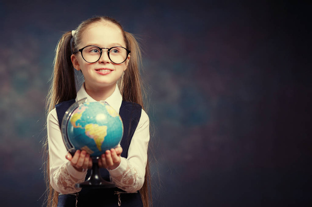 Блондинка маленька школярка тримає Світовий глобус в руці. Жінка-молодший студент починає вивчати географію. Гарненька дівчинка з двома довгими хвостами у шкільній уніформі Подивіться на модель карти Землі. Кольоровий тон
 - Фото, зображення