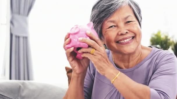 fröhliche asiatische Seniorin schüttelt Sparschwein lächelnd in die Kamera. Porträt einer reifen Frau, die ihr Sparschwein im Haus hält. 4k - Filmmaterial, Video