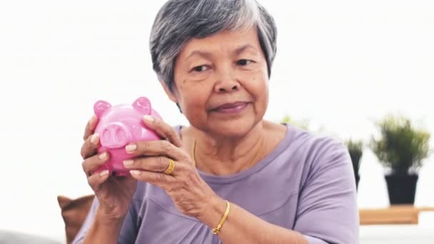 Cheerful asian senior woman shaking piggy bank smiling at camera. Portrait of mature woman saving money holding up piggy bank indoors. 4k - Footage, Video
