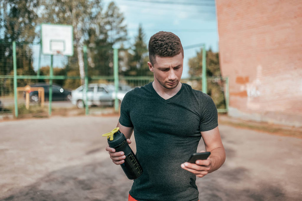 Male athlete, in summer sports field in hand smartphone, writes message to Internet, an app pulse and workout activity. Active lifestyle fitness workout. In his hand bottle of shaker with water. - Photo, image