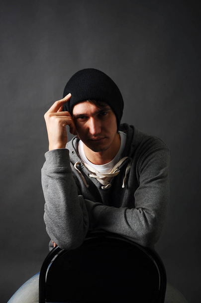 der junge Mann sitzt auf einem Stuhl in einem grauen Kapuzenpulli und einem schwarzen Hut, hält die Hand vor den Kopf im Schatten. - Foto, Bild