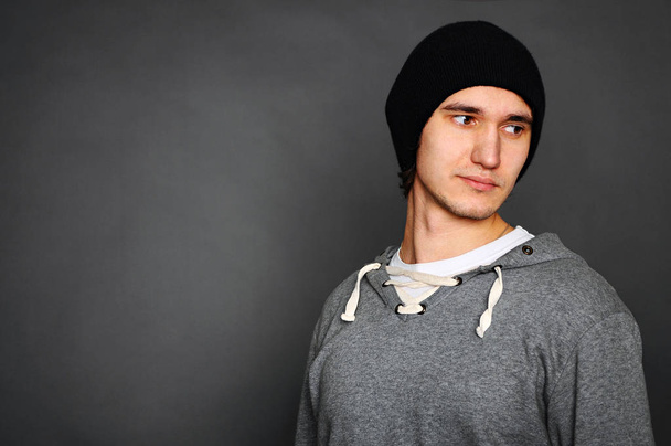 Portréja egy fiatalember egy szürke kapucnis pulcsit és egy fekete kalapot. Pózol egy szürke háttér. Stúdió felvétel. - Fotó, kép