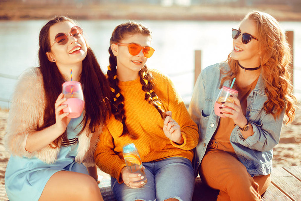 Three girls sit on the beach , near the river, talk, gossip, joke, laugh, smile at womans day. Girls on a hot day drink lemonade. Hipster model wearing sunglasses at summer day. Summer vibes. - Photo, Image