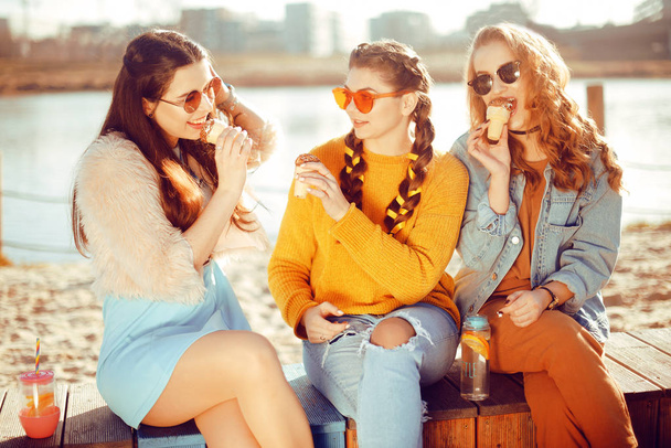 Drei Mädchen sitzen am Strand, in der Nähe des Flusses, reden, tratschen, scherzen, lachen, lächeln am Frauentag. Mädchen an einem heißen Tag essen Eis. Hipster-Model mit Sonnenbrille am Sommertag. Mädchen mit Frisur - Foto, Bild