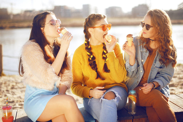 Троє дівчат сидять на пляжі, біля річки, розмовляють, пліткують, жартують, сміються, посміхаються в жіночий день. Дівчата в спекотний день їдять морозиво. Хіпстерська модель носить сонцезахисні окуляри в літній день. Дівчина з волоссям
 - Фото, зображення