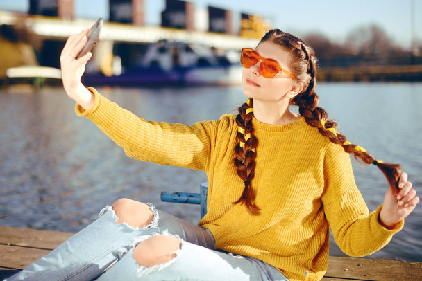 Beautiful girl sitting on the pier near the river. Hipster model yellow with brunette long hair. The girl at hot sunny day taking pictures on phone camera, Summer vibes. Girl take a selfie. Drink - Photo, Image