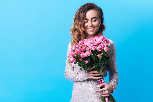 Young happy smiling redhead woman holding bouquet of colorful spring flowers isolated on blue background. Pink roses, festive bouquet in honor of women's day on March 8 or birthday - Fotoğraf, Görsel