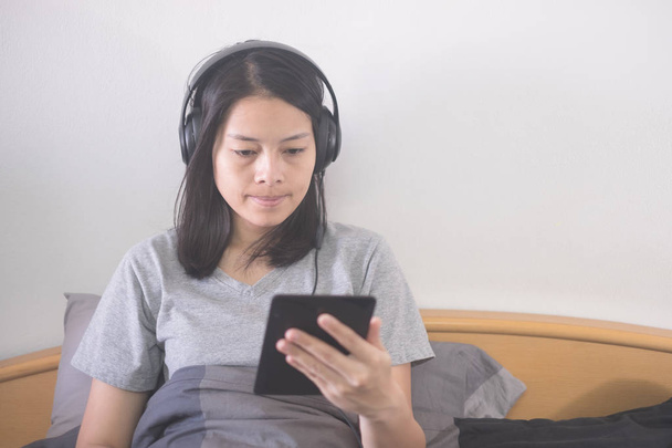 gyönyörű ázsiai nő hallgat zenét a fejhallgató és olvasó e betű-könyv az e-olvasók pihentető az ágyon - Fotó, kép