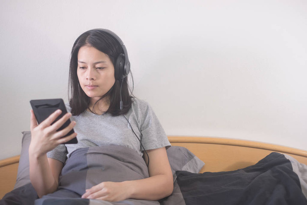 gyönyörű ázsiai nő hallgat zenét a fejhallgató és olvasó e betű-könyv az e-olvasók pihentető az ágyon - Fotó, kép